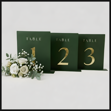 Three green table number signs with gold lettering, alongside a small floral arrangement.