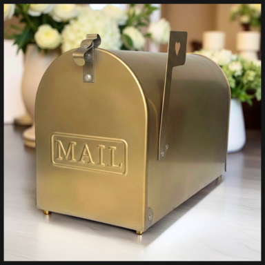Gold mailbox with the word "MAIL" displayed, placed on a surface with floral decorations.